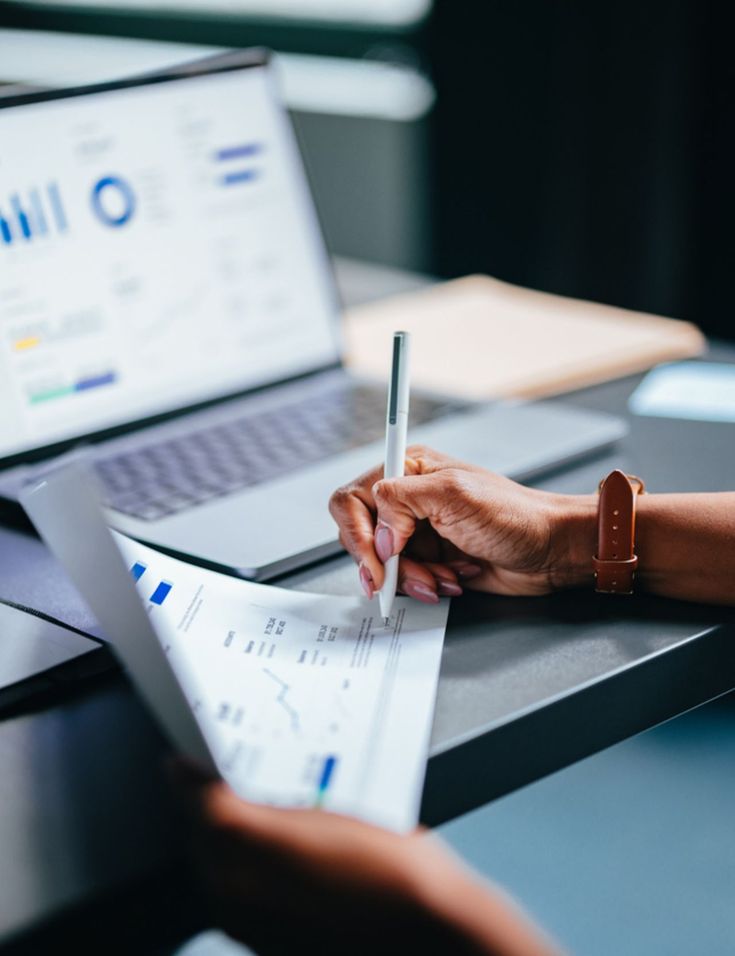 A hand signing a document beside a laptop showing financial charts